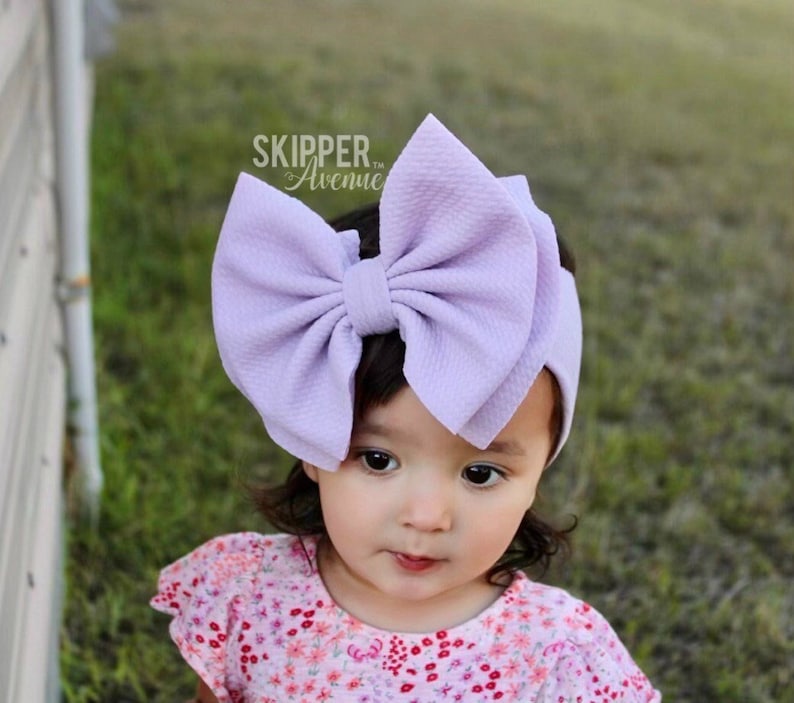 May include: A lavender bow headband with a large, fluffy bow. The headband is made of soft, stretchy fabric and is perfect for little girls.