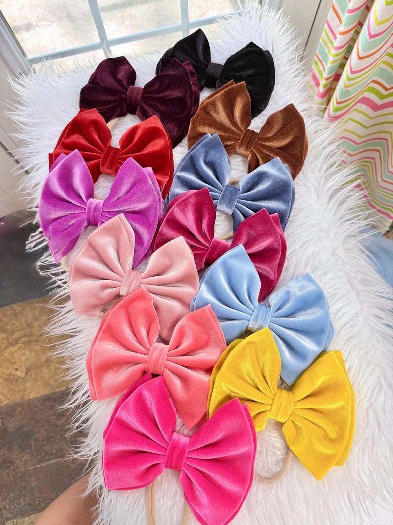 May include: A collection of velvet hair bows in various colors, including red, pink, blue, and yellow. The bows are arranged on a white, fluffy surface. Each bow is tied with a classic bow shape.