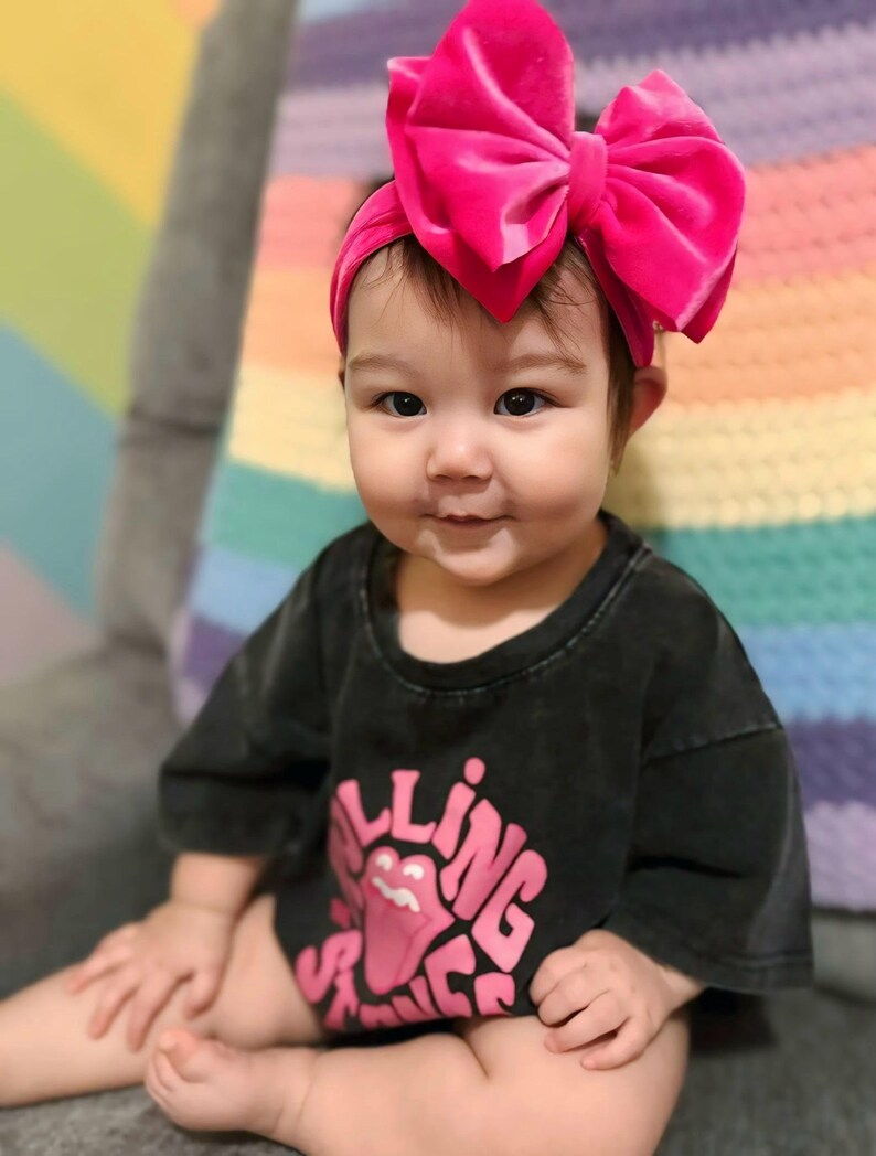 May include: A baby wearing a black t-shirt with the Rolling Stones logo in pink and a large pink velvet bow headband. The baby is sitting on a gray surface with a colorful blanket in the background.