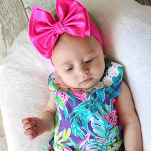 Puede incluir: Un beb&eacute; con una diadema de lazo rosa brillante y un vestido de estampado floral colorido. El beb&eacute; est&aacute; acostado sobre una almohada blanca.