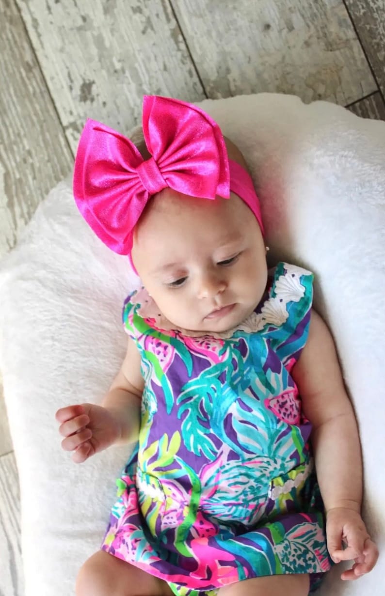 Puede incluir: Un beb&eacute; con una diadema de lazo rosa brillante y un vestido de estampado floral colorido. El beb&eacute; est&aacute; acostado sobre una almohada blanca.