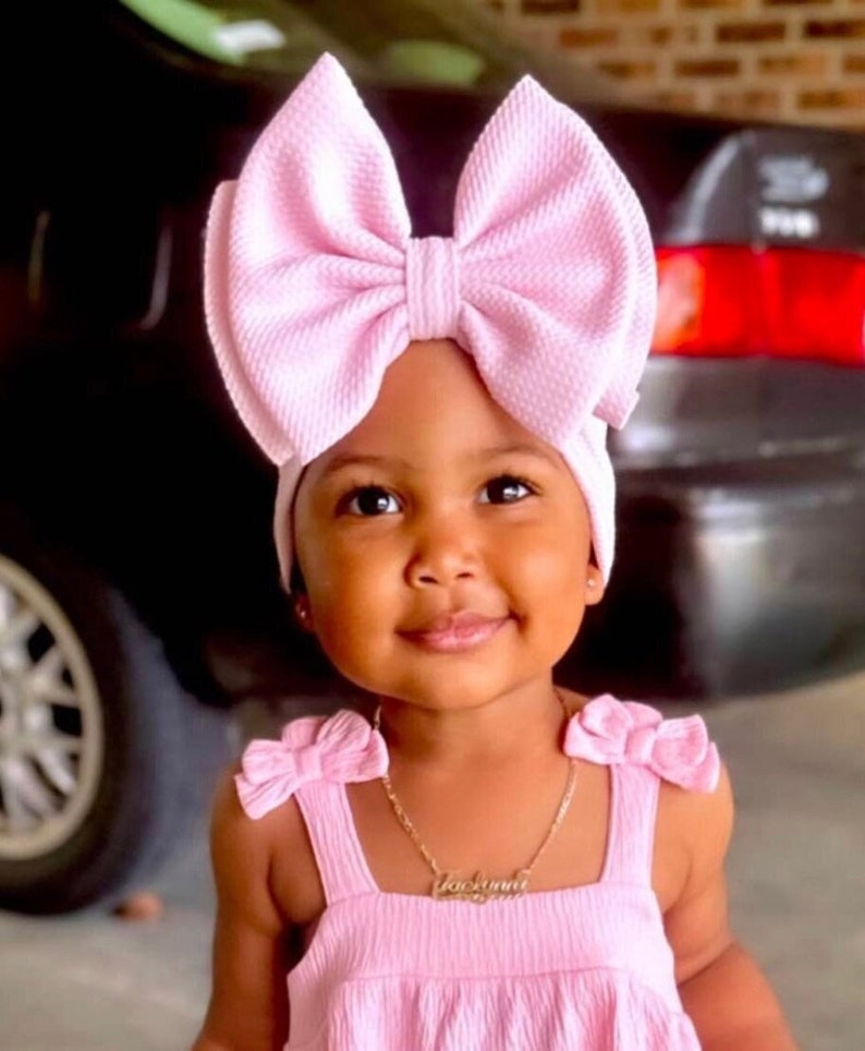 May include: A young child wearing a pink headband with a large bow and a pink dress with a gold necklace that says 'Jasmine'.