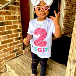 May include: A young child wearing a white t-shirt with a large pink glittery number 2 and the word "LEGIT" in colorful letters. The child is also wearing black ripped leggings, pink heart-shaped sunglasses, and a large white bow. The child is standing on wooden steps.