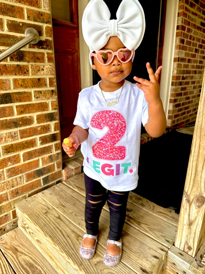 May include: A young child wearing a white t-shirt with a large pink glittery number 2 and the word "LEGIT" in colorful letters. The child is also wearing black ripped leggings, pink heart-shaped sunglasses, and a large white bow. The child is standing on wooden steps.