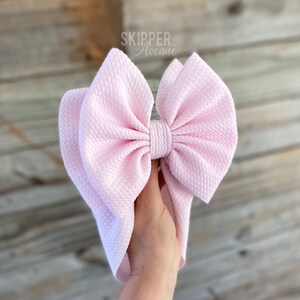 May include: A large pink bow made of textured fabric. The bow is tied in a knot and has a loop on the top. The bow is held in a hand.
