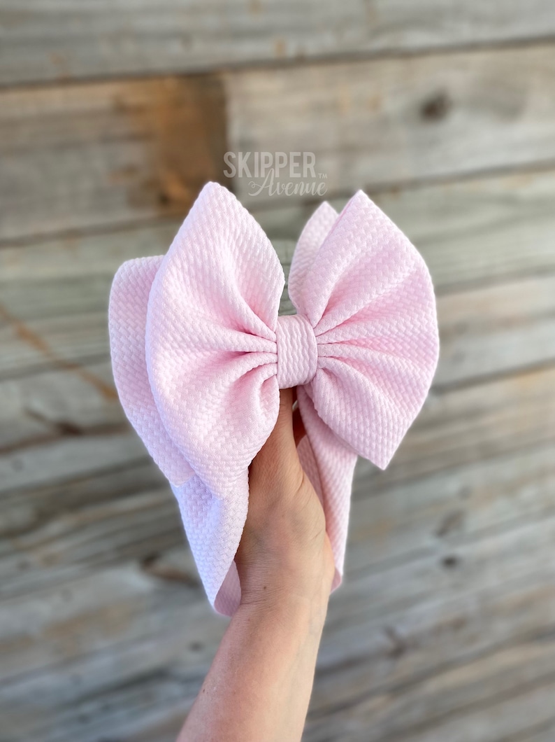 May include: A large pink bow made of textured fabric. The bow is tied in a knot and has a loop on the top. The bow is held in a hand.