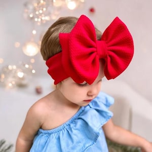 May include: A red fabric headband with a large, oversized bow. The headband is worn by a young child.