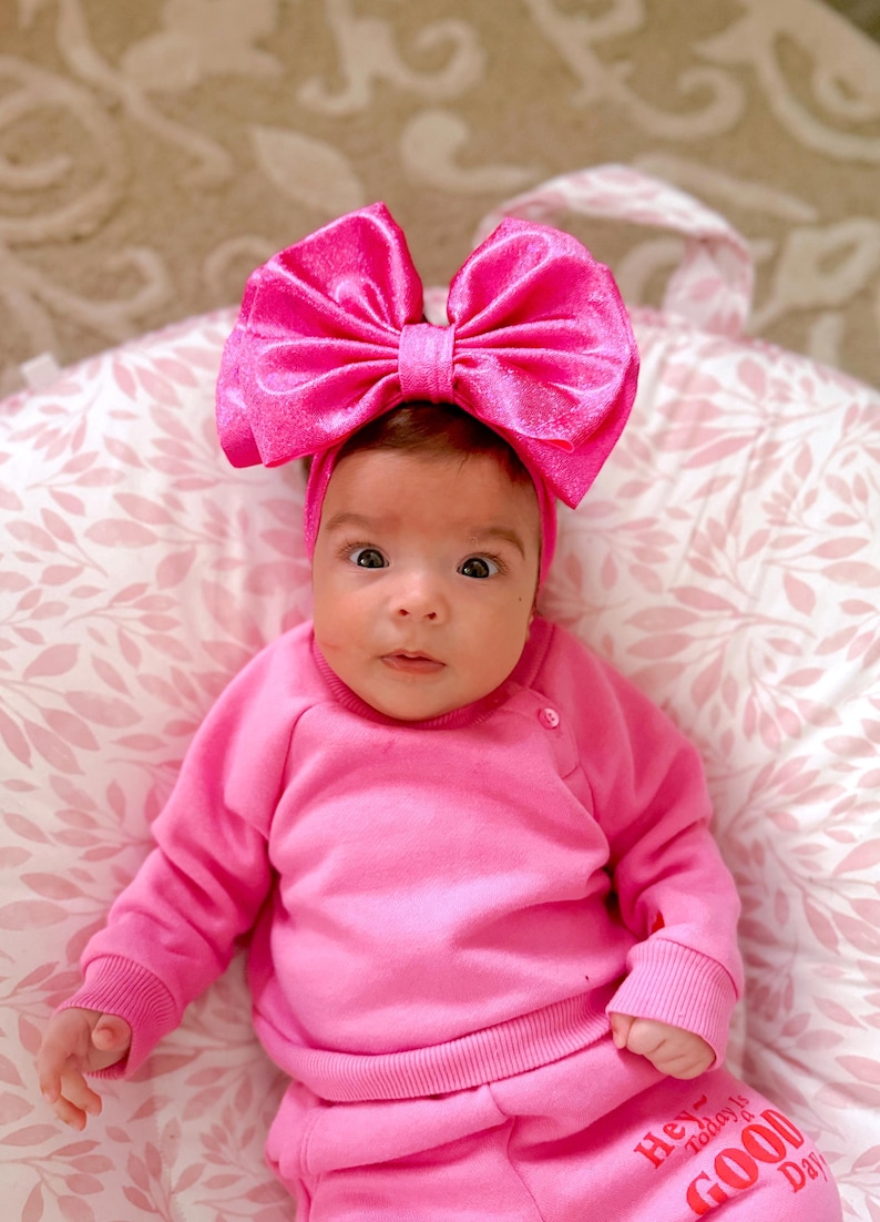 Puede incluir: Una ni&ntilde;a peque&ntilde;a con un jersey rosa y pantalones con el texto "Hey today is a GOOD Day" impreso en los pantalones. Lleva una diadema rosa grande con un lazo.