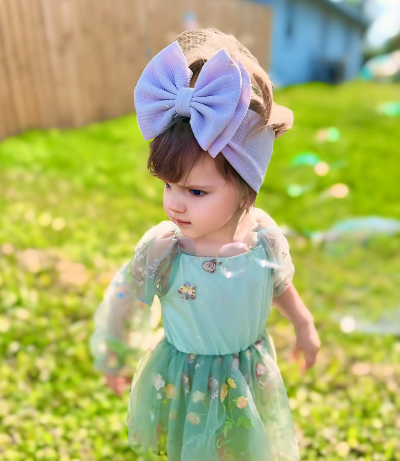 May include: A young girl wearing a light green dress with floral embroidery and a large lavender bow headband. The dress has sheer sleeves and a tulle skirt.
