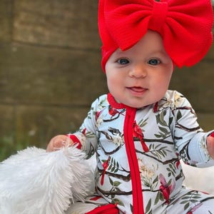 Op de afbeelding: Een baby draagt een blauwe en rode lange mouwen romper met een kardinaalvogel print. De romper heeft een rode rits en rode afwerking. De baby draagt een grote rode strik op zijn hoofd.