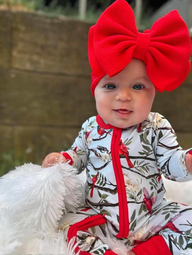 Op de afbeelding: Een baby draagt een blauwe en rode lange mouwen romper met een kardinaalvogel print. De romper heeft een rode rits en rode afwerking. De baby draagt een grote rode strik op zijn hoofd.