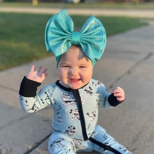 Puede incluir: Un beb&eacute; con un mono azul y negro con estampado de Mickey Mouse y un gran lazo de purpurina turquesa en la cabeza. El beb&eacute; est&aacute; sentado en una acera y sonriendo.