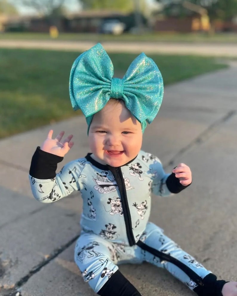 Puede incluir: Un beb&eacute; con un mono azul y negro con estampado de Mickey Mouse y un gran lazo de purpurina turquesa en la cabeza. El beb&eacute; est&aacute; sentado en una acera y sonriendo.