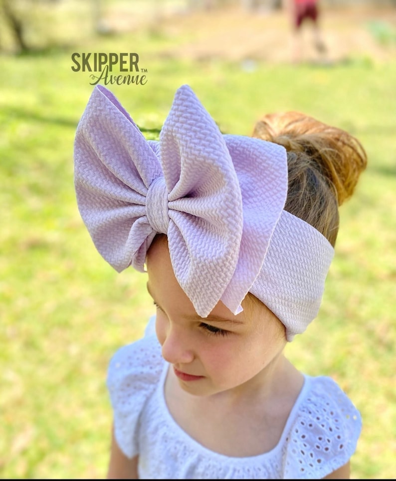 May include: A large, lavender-colored bow headband. The bow is made of a textured fabric and is tied in a large, floppy knot. The headband is worn by a young girl with brown hair.