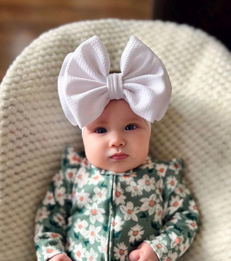May include: A baby wearing a white bow headband and a green floral print onesie. The baby is lying on a cream-colored, textured surface. The onesie has a zipper closure and ruffle details.