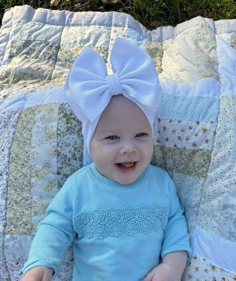 May include: A smiling baby wearing a light blue long-sleeved shirt with a decorative lace trim and a large white bow headband. The baby is lying on a patchwork quilt with floral and geometric patterns.