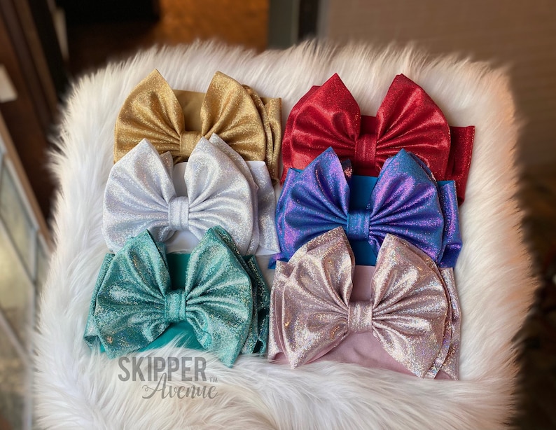 May include: Six sparkly fabric headbands with large bows in various colors including gold, white, teal, purple, pink, and red. The headbands are arranged in two rows of three. The bottom row has the words 'SKIPPER Avenue™' printed on the fabric.
