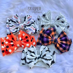 May include: A selection of Halloween-themed baby headbands. The headbands display patterns such as bats, spiderwebs, polka dots, and plaid. The colour scheme includes orange, black, white, and purple. The brand name "Skipper Avenue" is visible.
