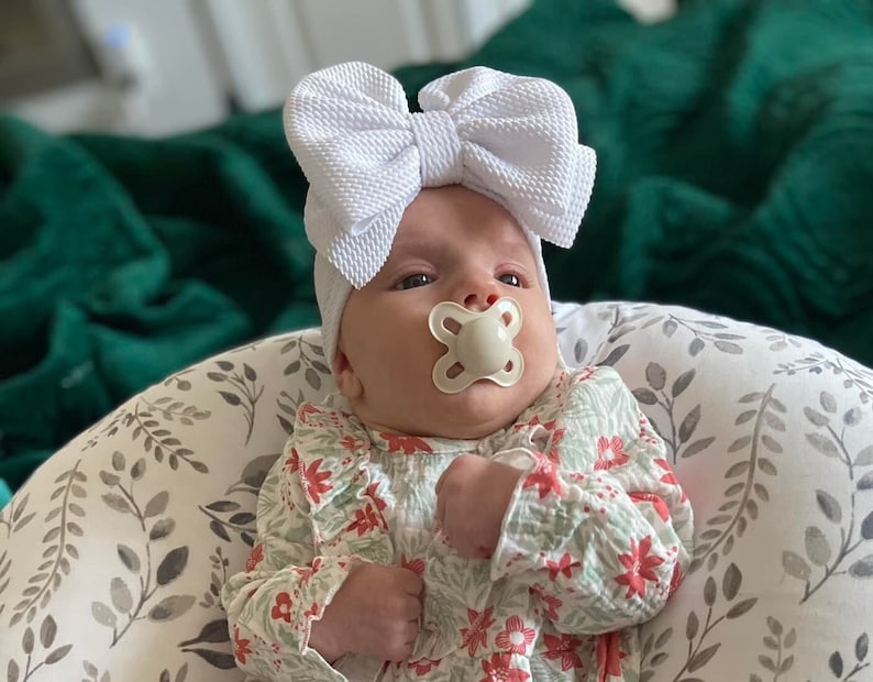 May include: A baby wearing a white bow headband and a floral print onesie. The baby is using a pacifier and is seated in a patterned baby lounger. The background includes a green blanket.