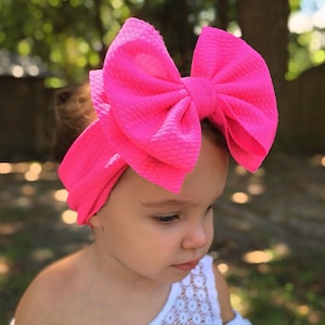 May include: A bright pink baby headband with a large bow. The bow is textured and sits atop a matching pink band. The background is blurred, with green foliage and sunlight.