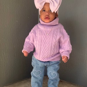 May include: A young child wearing a lavender cable knit sweater with a turtleneck and a large lavender bow headband. The child is also wearing light blue jeans with white heart embroidery and white sneakers.