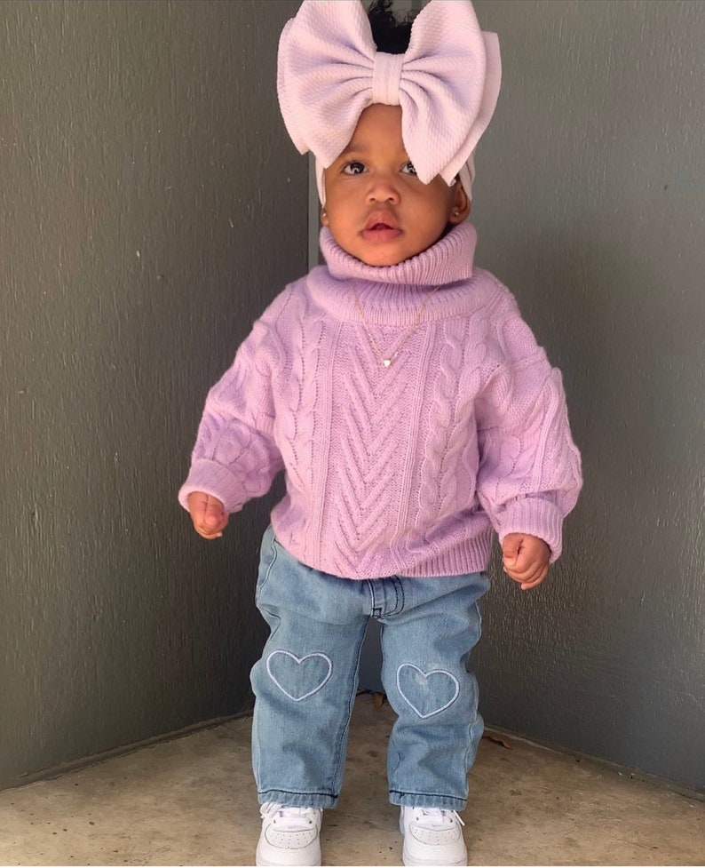 May include: A young child wearing a lavender cable knit sweater with a turtleneck and a large lavender bow headband. The child is also wearing light blue jeans with white heart embroidery and white sneakers.