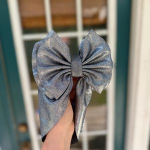 May include: A large, silver bow headband with a shimmering, textured finish. The bow is centered and has a classic bow shape. The headband is a stylish hair accessory.