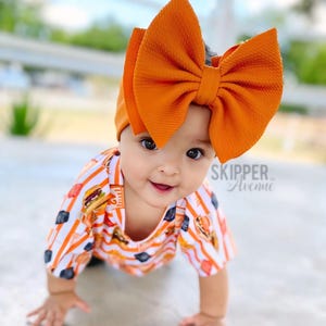 May include: A baby wearing a white and orange striped shirt with a large orange bow headband. The shirt has a pattern of burgers and fries. The baby is crawling on a concrete surface. "SKIPPER Avenue" is written on the shirt.