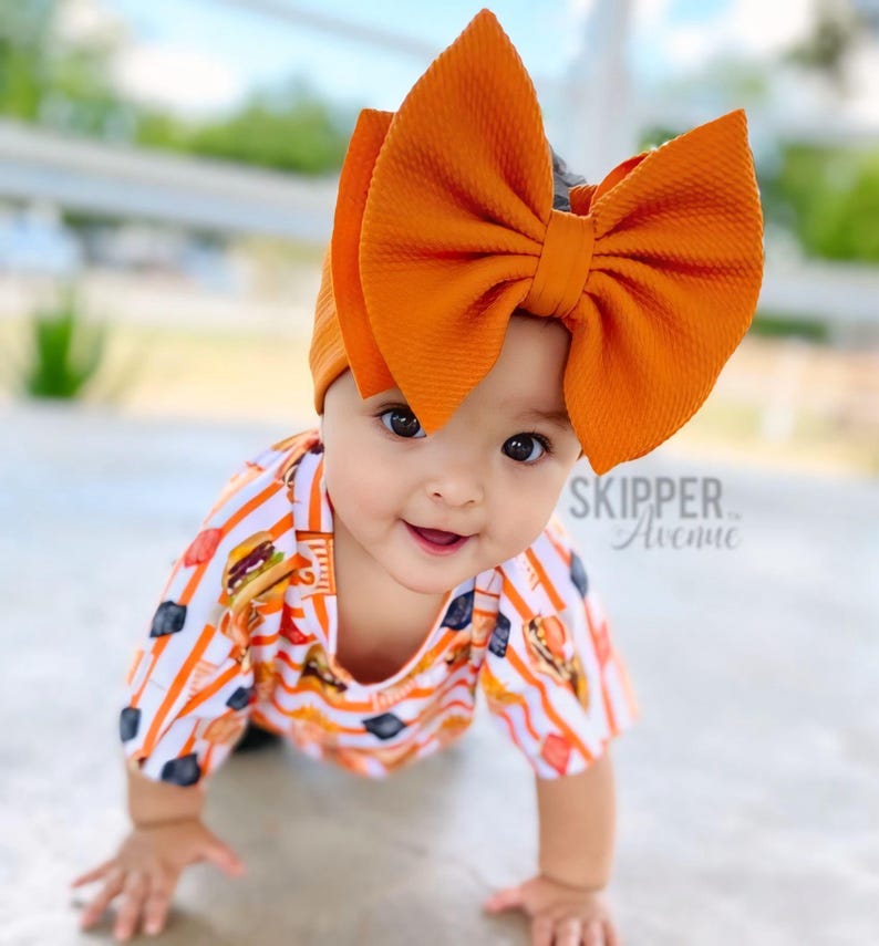 May include: A baby wearing a white and orange striped shirt with a large orange bow headband. The shirt has a pattern of burgers and fries. The baby is crawling on a concrete surface. "SKIPPER Avenue" is written on the shirt.