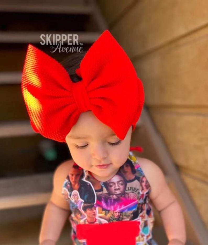 Op de afbeelding: Een rood strik hoofdband op het hoofd van een kind. Het hoofdband is gemaakt van een zachte, textuurrijke stof. Het kind draagt een kleurrijk shirt met een Stranger Things-thema.