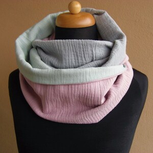 May include: A soft, cozy infinity scarf in a three-tone color scheme. The scarf is made of a textured fabric and features a light gray, light green, and pink color combination.