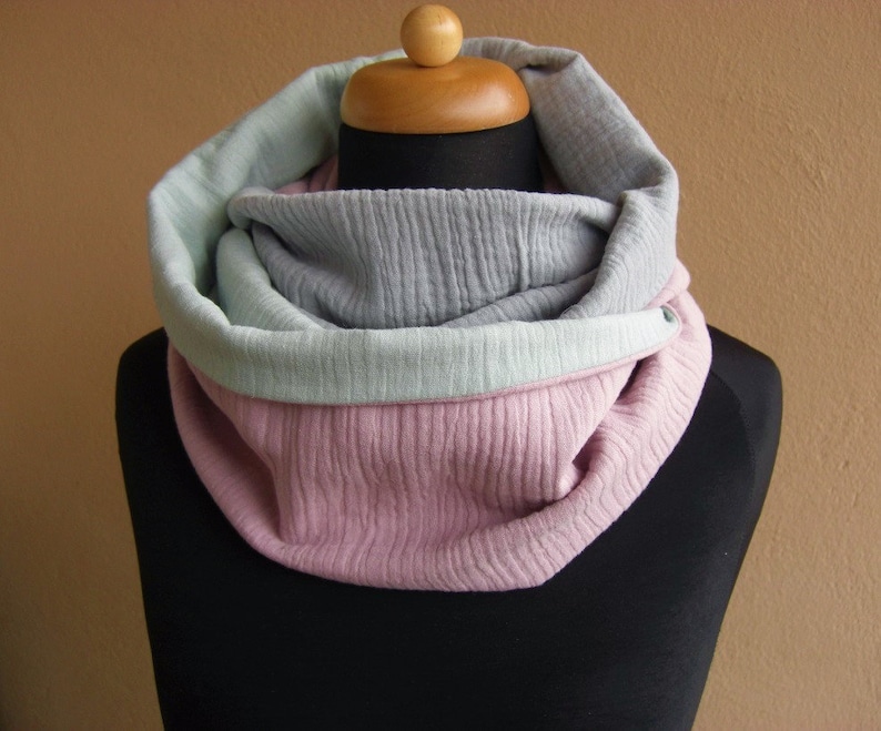 May include: A soft, cozy infinity scarf in a three-tone color scheme. The scarf is made of a textured fabric and features a light gray, light green, and pink color combination.