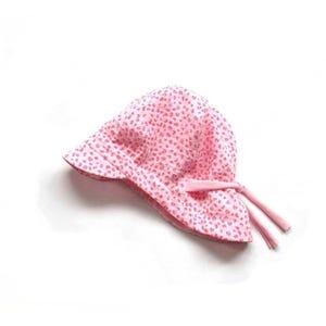 Könnte beinhalten: Ein rosa Sonnenhut mit einem kleinen, sich wiederholenden Muster aus dunkleren rosa Punkten. Der Hut hat eine Krempe und Bindebänder auf beiden Seiten. Der Hut ist aus einem weichen Material gefertigt.