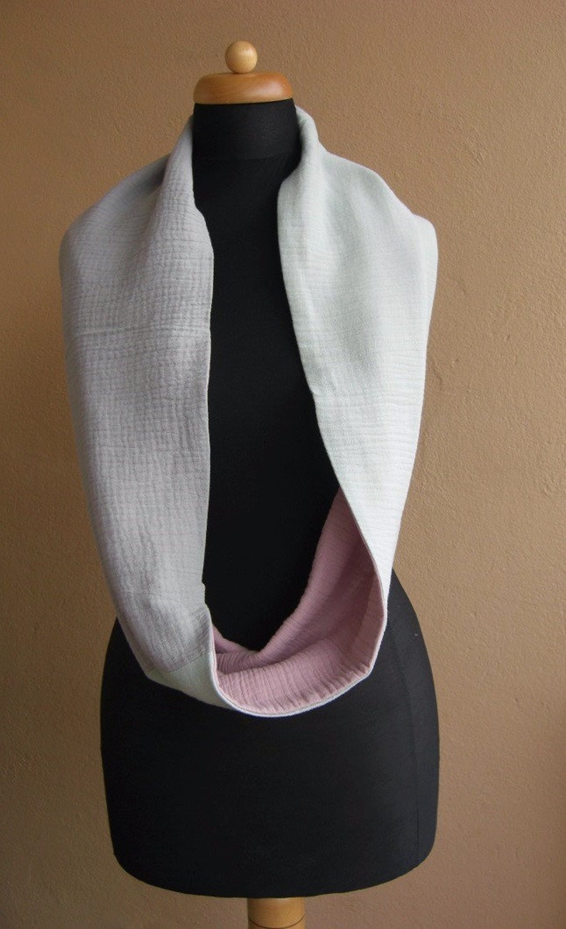 May include: A light gray infinity scarf with a pink lining. The scarf is displayed on a black mannequin.