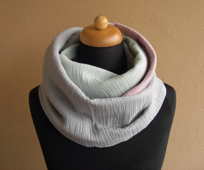 May include: A soft, gray infinity scarf with a pink and light green ombre effect. The scarf is made of a textured fabric and is folded in a loop.