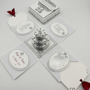 Könnte beinhalten: Eine weiße und silberne Explosionsbox mit einem silbernen Kuchen, einem Paar und dem Text "25 Jahre glücklich vereint" und "Marga & Thomas 18. Oktober 1999" in silberner Schrift.