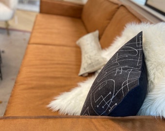 Könnte beinhalten: Ein langes, hellbraunes Sofa mit glatter Oberfläche, ausgestattet mit einem dunkel anthrazitfarbenen Kissen mit weißen Linien, einem weißen Kunstfellkissen und einem kleinen rechteckigen hellgrauen Kissen mit Gittermuster.