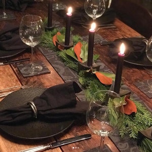 K&ouml;nnte beinhalten: Ein gedeckter Tisch mit schwarzen Stabkerzen in dunklen Metallhaltern, arrangiert mit gr&uuml;nen Kiefernnadeln und orangefarbenen Bl&auml;ttern. Das Gedeck umfasst schwarze Teller, schwarze Stoffservietten mit silbernen Ringen, klare Weingl&auml;ser und silbernes Besteck.