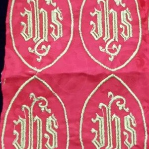 May include: Red fabric with four embroidered oval designs. Each oval features the gold letters "IHS" in a stylized font, surrounded by a gold border. The fabric has a subtle pattern.