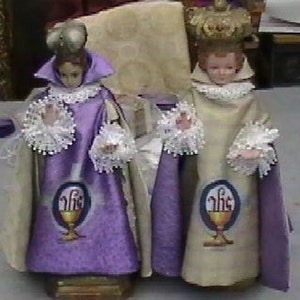 May include: Two small, purple and white robed figurines of children wearing crowns. The figurines are standing on a wooden base. The robes have a chalice and host design.