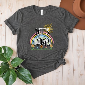 May include: Dark grey t-shirt featuring the text "Let's Learn Today" above a rainbow and flowers. A yellow sun is in the upper right. The shirt is folded and tied at the bottom. A brown hat is in the background.
