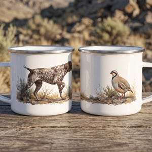 May include: Two white enamel mugs with silver rims. The left mug features a brown and white spotted hunting dog standing in grass. The right mug displays a small brown, grey, and red bird standing on rocky ground. Both mugs have side handles.