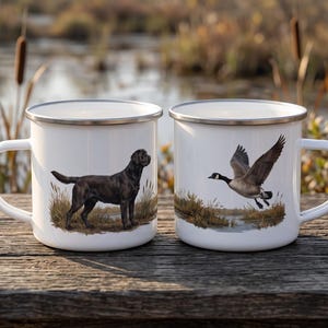 Może przedstawiać: Dwa białe emaliowane kubki ze srebrnymi obwódkami. Na lewym kubku znajduje się czarny labrador retriever stojący w wysokiej trawie. Na prawym kubku widnieje gęś kanadyjska w locie nad bagnistym krajobrazem. Oba kubki posiadają uchwyt.