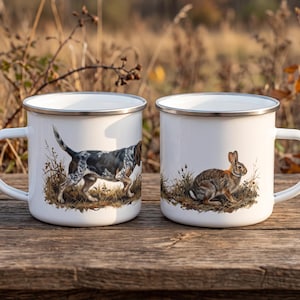 Puede incluir: Dos tazas de esmalte blanco con bordes plateados. La taza de la izquierda presenta una ilustración detallada de un perro de caza moteado en un campo de hierba. La taza de la derecha muestra una ilustración detallada de un conejo marrón.