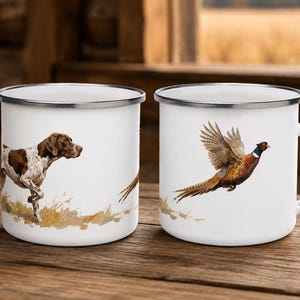 Puede incluir: Dos tazas de esmalte blanco con bordes plateados. Una taza muestra un perro de caza manchado de marrón y blanco en hierba alta. La otra taza muestra un faisán colorido en vuelo con plumas marrones, doradas y rojas sobre un parche de hierba.