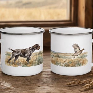 Puede incluir: Dos tazas de esmalte blanco con bordes y asas plateados. Una taza presenta un perro de muestra alemán de pelo corto marrón y blanco en un campo. La otra taza muestra un ave de caza marrón y blanca en vuelo sobre un paisaje herboso.