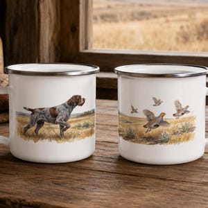 Könnte beinhalten: Zwei weiße Emaille-Tassen mit silbernen Rändern. Die linke Tasse zeigt einen braun-weißen Jagdhund, der in einem Feld steht. Die rechte Tasse zeigt eine Szene mit drei fliegenden Vögeln über einer grasbewachsenen Landschaft.