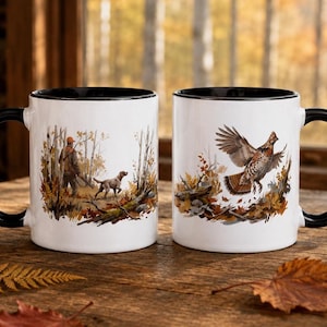 Könnte beinhalten: Zwei weiße Keramikbecher mit schwarzen Henkeln und Rändern. Ein Becher zeigt einen Jäger mit Hund im Wald. Der andere Becher zeigt ein Kragenhuhn im Flug über herbstlichem Laub. Beide Becher sind mit detaillierten, farbigen Illustrationen versehen.