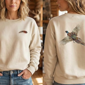 May include: Beige sweatshirt with a small, detailed insect design on the front and a pheasant in flight on the back. The pheasant has brown, green, and blue feathers. The sweatshirt is a neutral color.