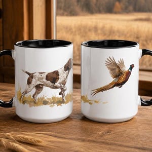May include: Two white ceramic mugs with black interiors and black handles. The left mug features a brown and white spotted hunting dog standing in grass. The right mug displays a colourful pheasant in flight above a patch of dry grass.